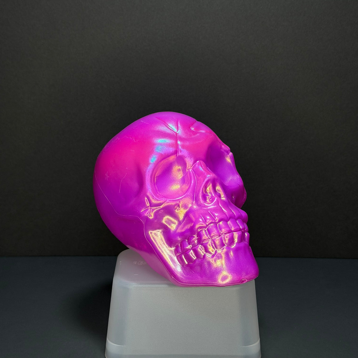 A glossy, iridescent pink skull sculpture displayed on a translucent white pedestal against a dark gray background.