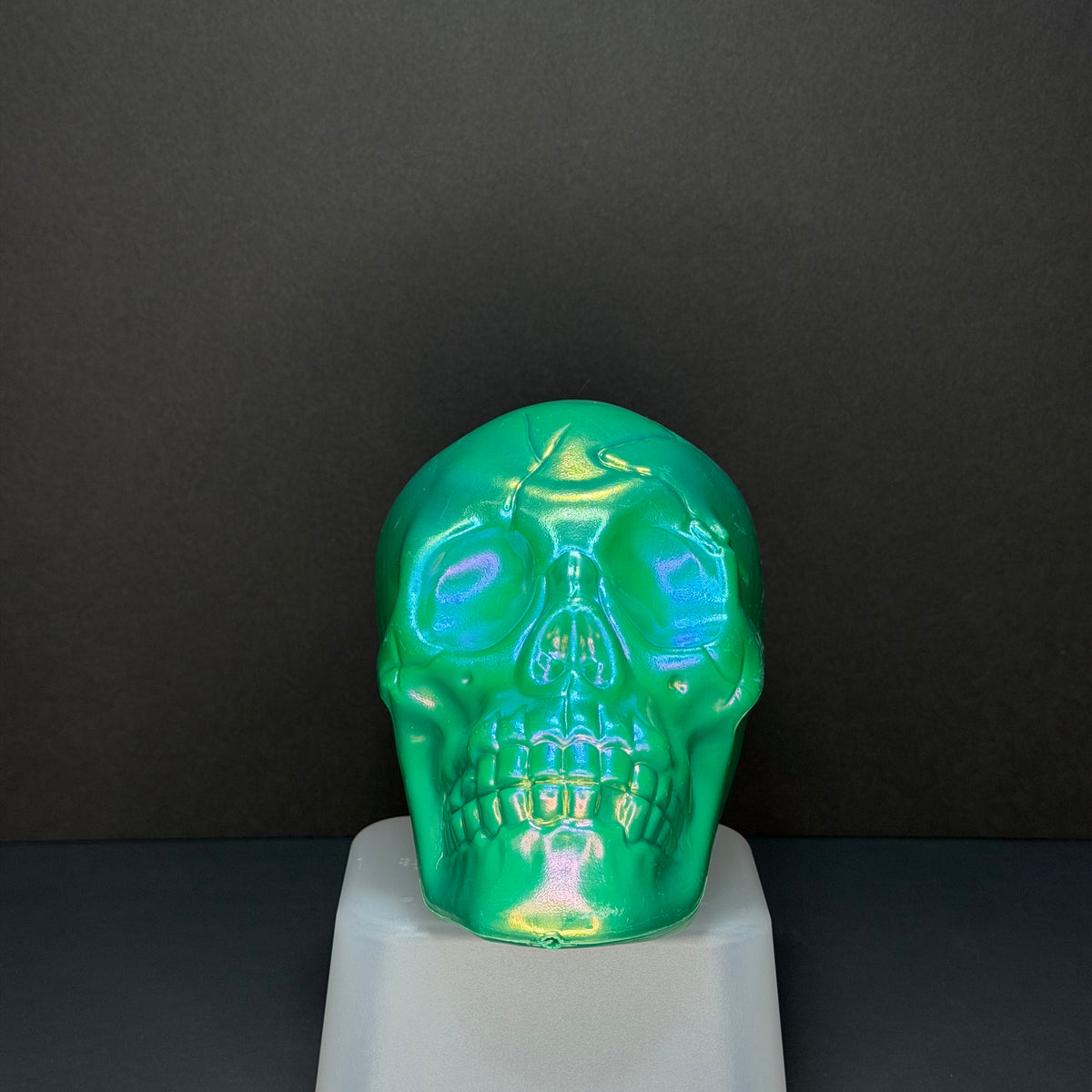 A glossy, iridescent green skull-shaped object displayed on a white pedestal against a dark gray background.