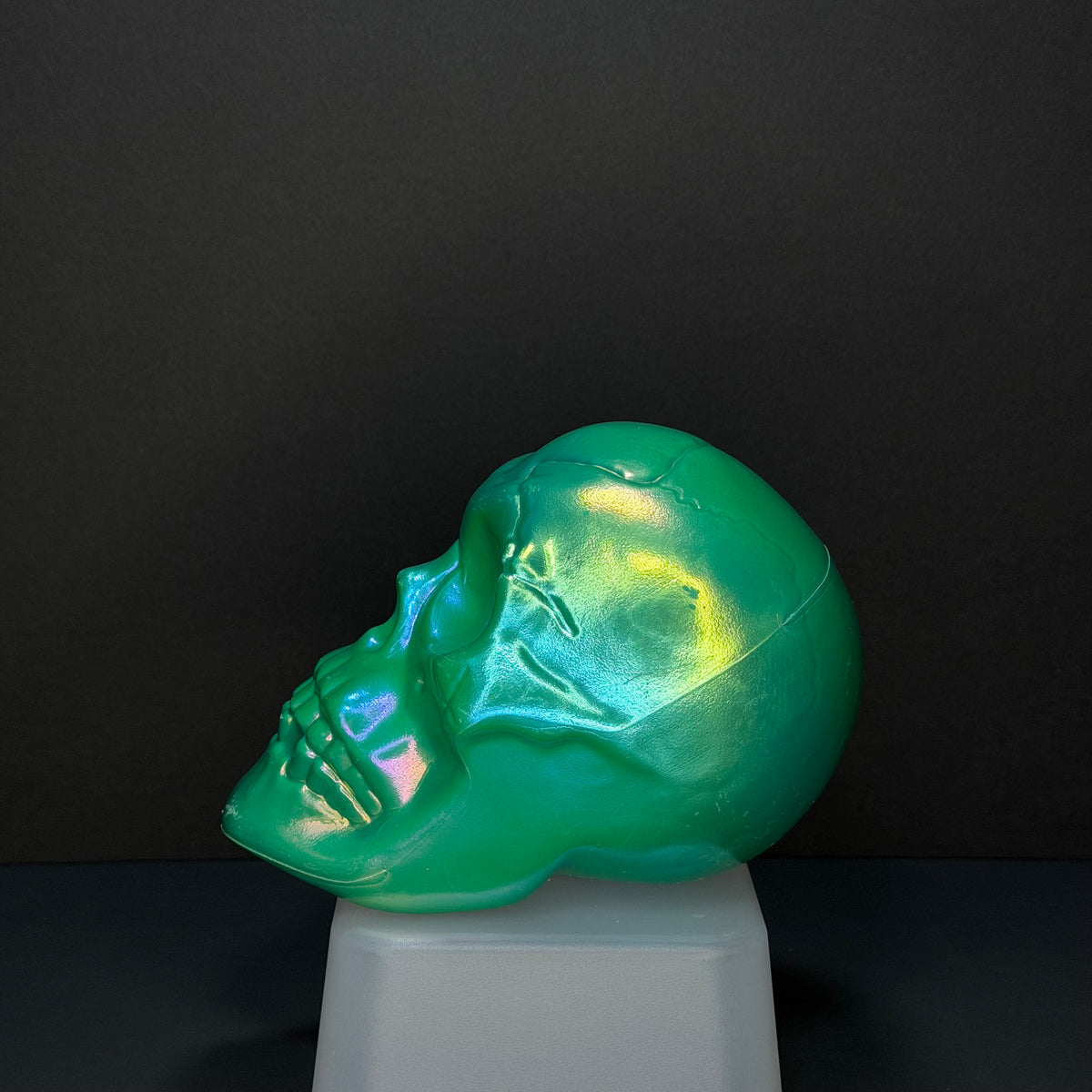 An iridescent green skull sculpture displayed on a white pedestal against a dark gray background.