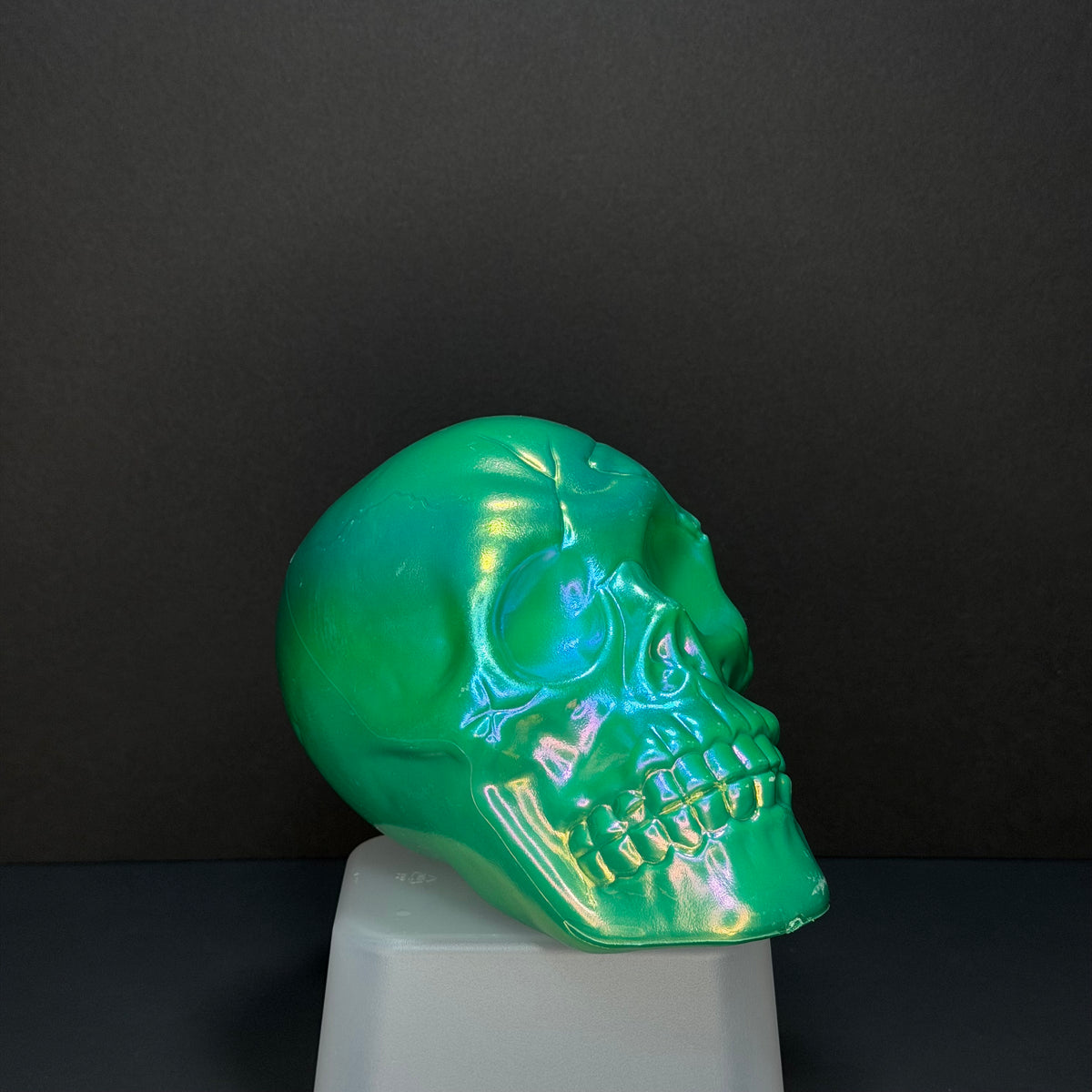 A glossy, iridescent green skull sculpture displayed on a white pedestal against a dark gray background.