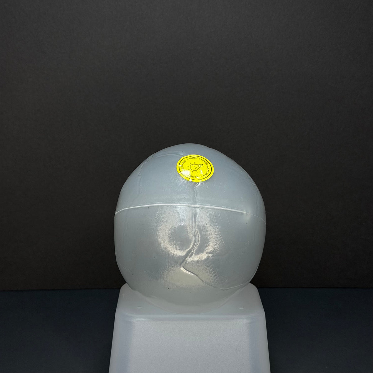 A translucent white plastic spherical object with a small yellow circular sticker on top, displayed on a white pedestal against a dark gray background. Glow in the dark