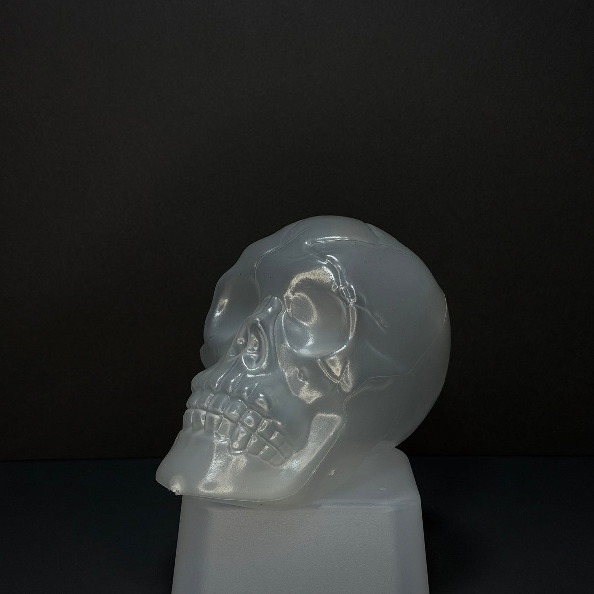 A translucent frosted glass or acrylic skull sculpture displayed on a matching pedestal against a dark black background. Glow-in-the-dark