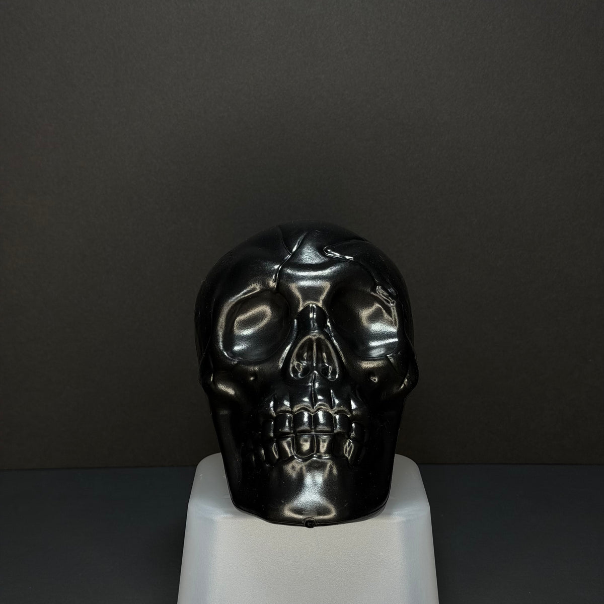 A glossy black skull sculpture displayed on a white pedestal against a dark gray background.