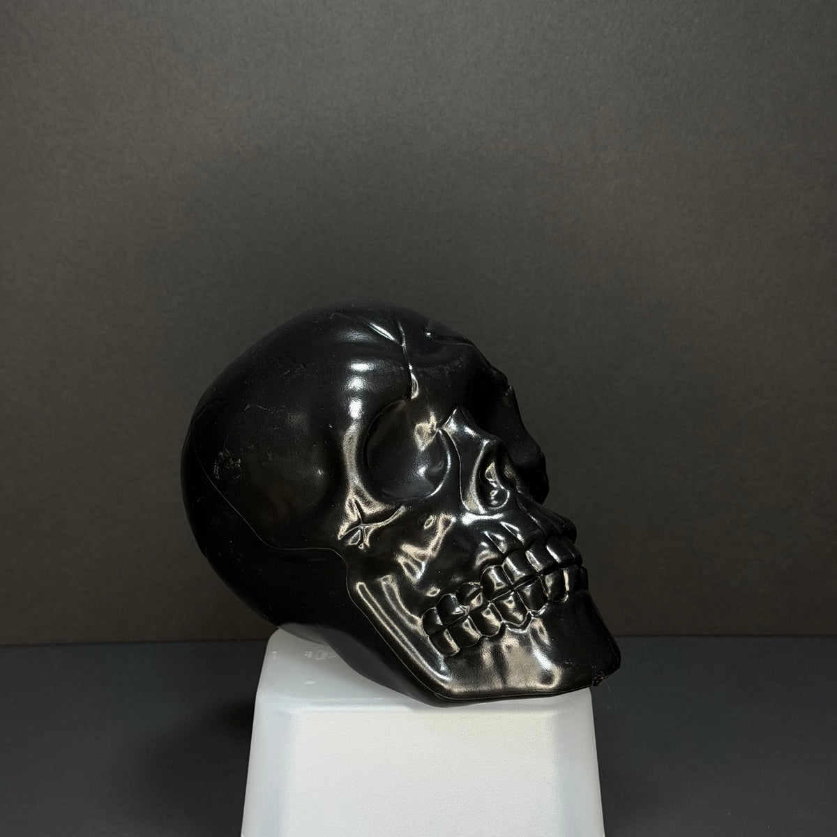 A glossy black skull sculpture displayed on a white pedestal against a dark gray background.