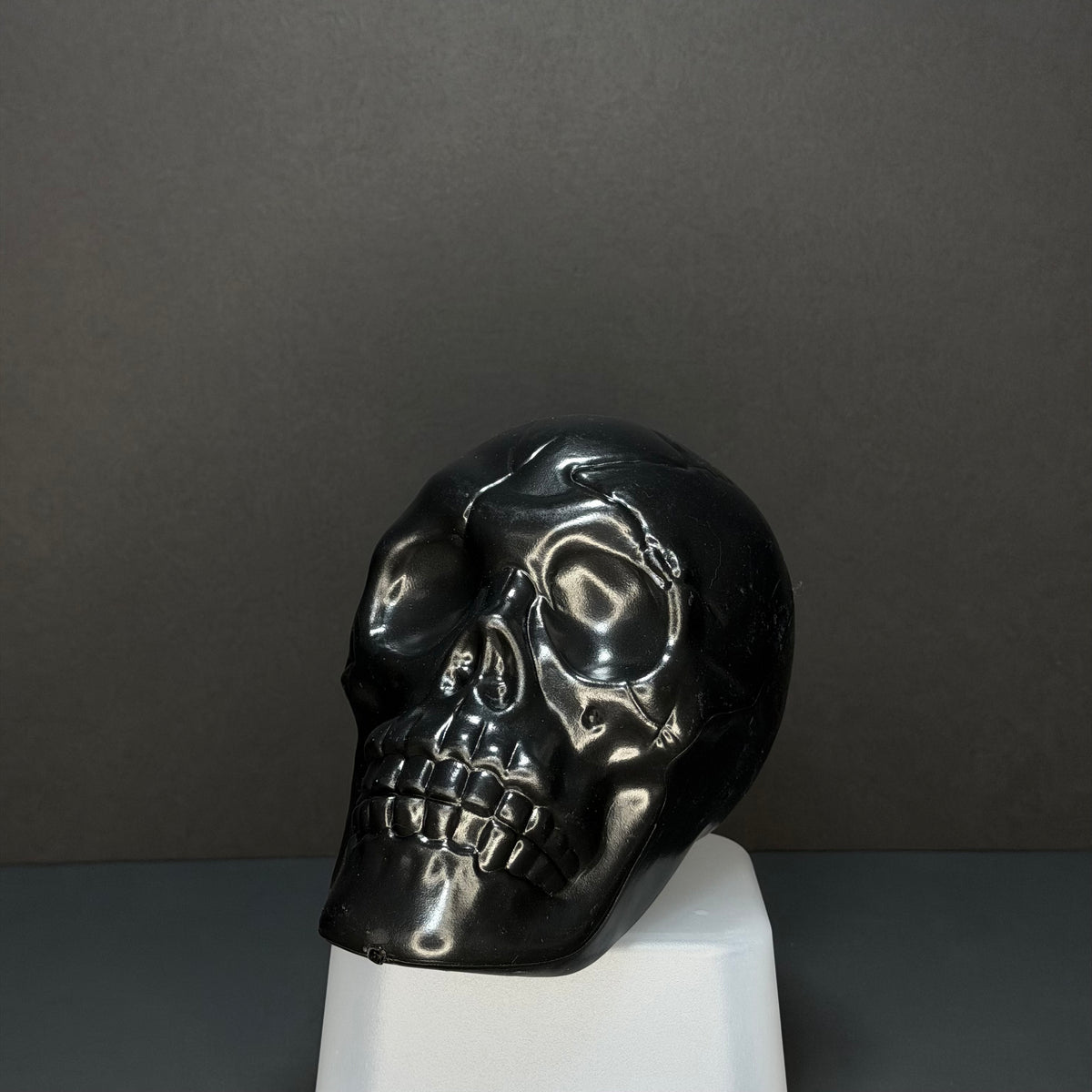 A glossy black polished skull sculpture displayed on a white pedestal against a dark gray background.
