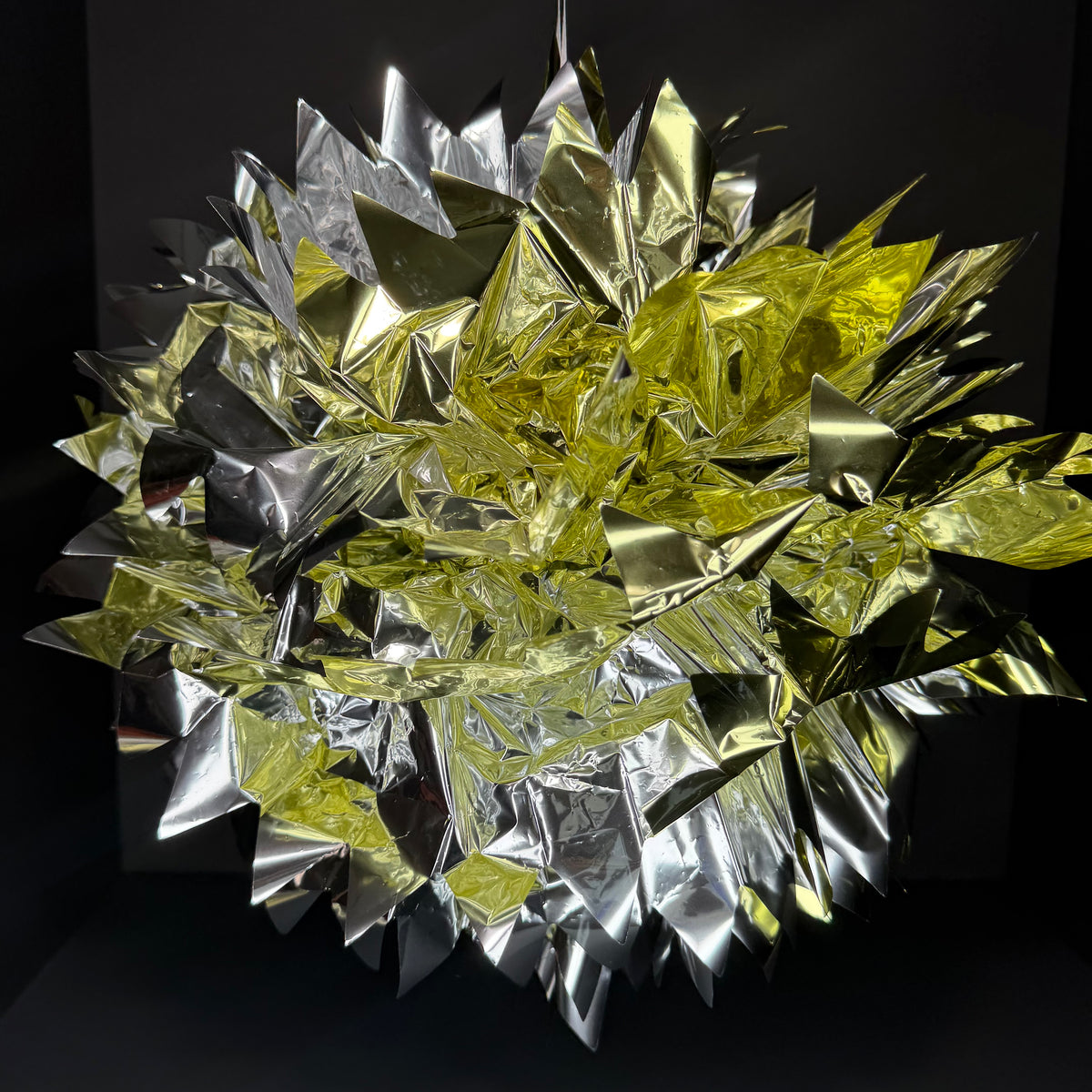 A metallic, faceted decorative object made of overlapping reflective foil-like panels in silver and yellow-gold tones, photographed against a dark studio background.