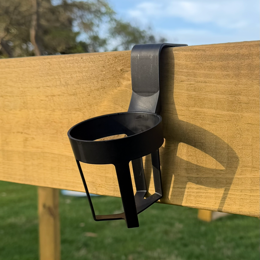 A black metal cup holder or small cup mount attached to a wooden fence outdoors, with grass and trees in the background.